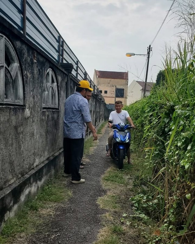 Seorang penunggang motosikal melalui laluan sempit di antara tembok dan tumbuhan.