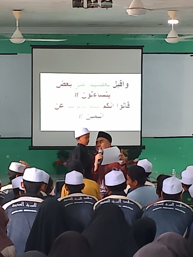 Seorang pembimbing menggunakan mikrofon bersama seorang pelajar di hadapan dewan.